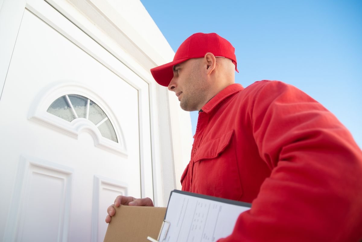 Un livreur en uniforme rouge tenant un colis devant une porte blanche, prêt à